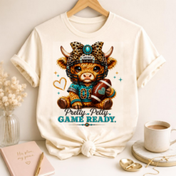 Pretty Petty Game Ready Highland Cow football design displayed on a white t-shirt mockup with leopard bow, football helmet, and turquoise accents.